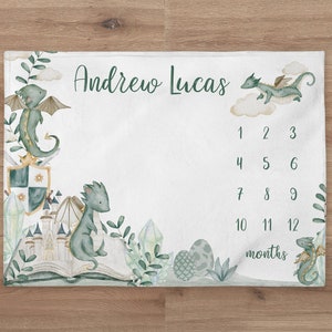 May include: A white baby milestone blanket with a green dragon and castle design. The blanket features the name "Andrew Lucas" and a chart to track the baby's growth from 1 to 12 months.