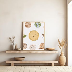 K&ouml;nnte beinhalten: Ein wei&szlig;es Poster mit einer Cartoon-Illustration einer gelben Sonne und Planeten mit l&auml;chelnden Gesichtern. Die Planeten sind braun, gr&uuml;n, blau und orange. Das Poster ist in einem d&uuml;nnen braunen Rahmen gerahmt und h&auml;ngt an einer wei&szlig;en Wand.