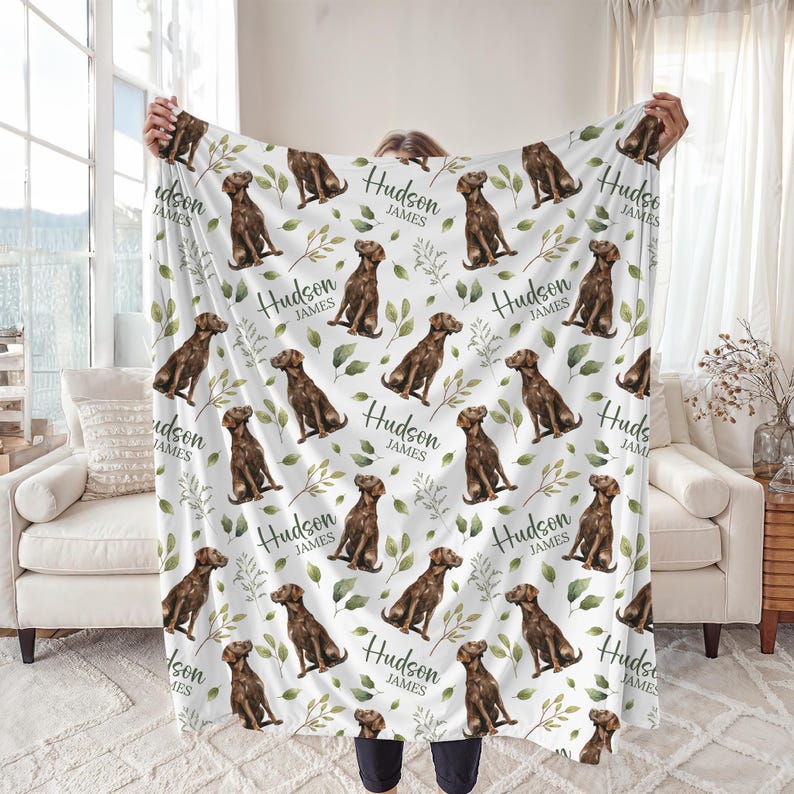 May include: A white blanket featuring a repeating pattern of brown dogs, green leaves, and the name "Hudson James" in black script. The blanket is held up, showcasing the design and soft texture.