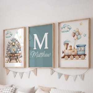 May include: Three framed watercolor prints with a train theme. One print features a train, another has the letter "M" and the name "Matthew", and the third shows a train with balloons. The frames are light wood, and the background is white.