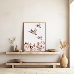 Peut inclure: Une impression d'art encadr&eacute;e repr&eacute;sentant trois canards en vol sur fond blanc. Le bas de l'impression pr&eacute;sente un arrangement floral dans les tons de rose, marron et blanc. Le cadre est de couleur marron clair.