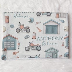 May include: A light-colored baby blanket with a repeating pattern of motorcycles, garages, helmets, and tools in shades of orange, blue, and gray. The names "Robinson" and "Anthony Robinson" are written in script.
