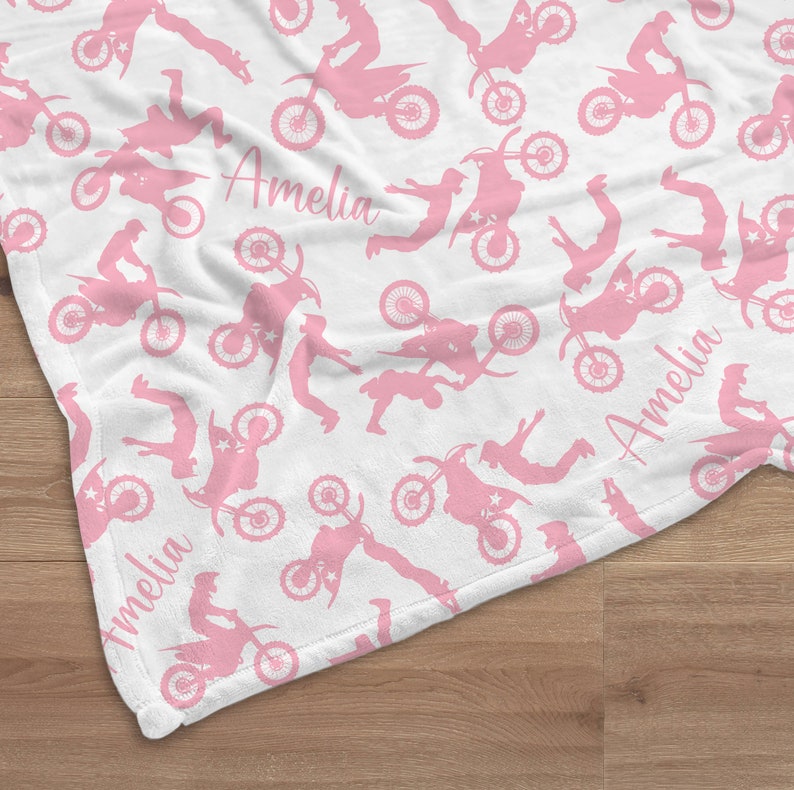 May include: A white fleece blanket with a pink repeating pattern of motocross riders. The blanket has the name "Amelia" written three times in pink.