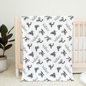 K&ouml;nnte beinhalten: Wei&szlig;e Babydecke mit einem sich wiederholenden Muster aus grauen Motorradillustrationen und dem Namen "Wallace". Die Decke ist &uuml;ber ein Holzbettchen drapiert.