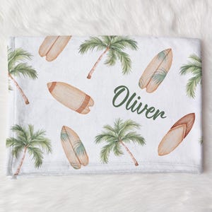 May include: White baby blanket with a tropical theme. The blanket features a repeating pattern of palm trees and surfboards. The name "Oliver" is written in green cursive lettering.