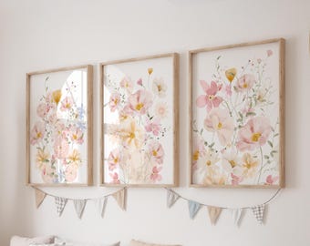 May include: Three framed floral art prints featuring watercolor paintings of pink, yellow, and white flowers. The prints have light brown wooden frames and are hung on a white wall. A decorative banner hangs below the prints.