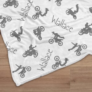 K&ouml;nnte beinhalten: Wei&szlig;e Decke mit einem sich wiederholenden Muster aus grauen Motocross-Fahrern in Aktion. Der Name "Wallace" ist in grauer Schrift &uuml;ber die Decke gedruckt. Die Decke liegt auf einer Holzoberfl&auml;che.