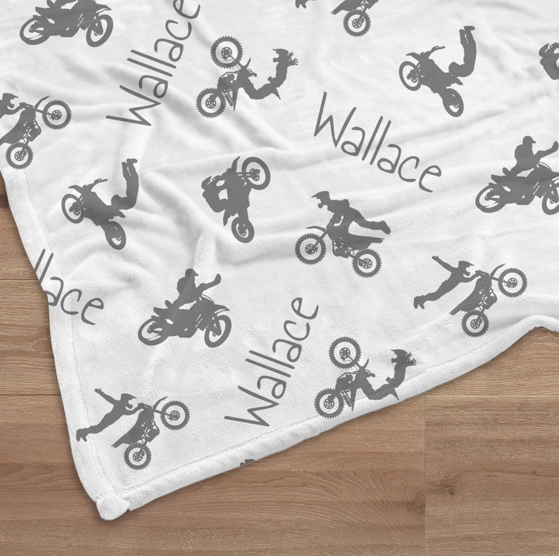 K&ouml;nnte beinhalten: Wei&szlig;e Decke mit einem sich wiederholenden Muster aus grauen Motocross-Fahrern in Aktion. Der Name "Wallace" ist in grauer Schrift &uuml;ber die Decke gedruckt. Die Decke liegt auf einer Holzoberfl&auml;che.