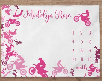 May include: Pink and white baby milestone blanket with a motocross theme. The blanket features pink silhouettes of motocross riders and motorcycles. The text "Madelyn Rose" is printed in pink cursive script. The numbers 1 through 12 are printed in black, along with the word "months".