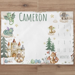 May include: A white baby milestone blanket with a green, brown, and gold castle, dragons, and stars. The blanket has the name "CAMERON" printed on it and a number chart for tracking a baby's growth from 1 to 12 months.