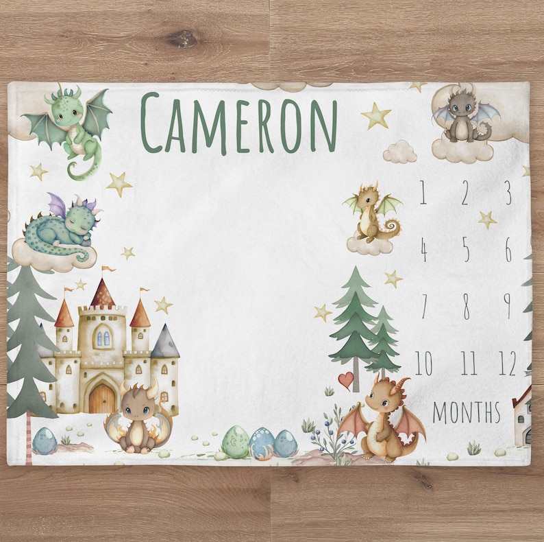 May include: A white baby milestone blanket with a green, brown, and gold castle, dragons, and stars. The blanket has the name "CAMERON" printed on it and a number chart for tracking a baby's growth from 1 to 12 months.