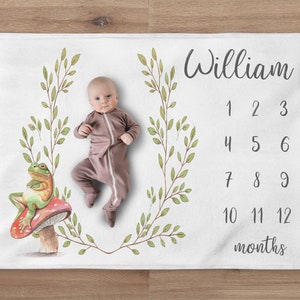 May include: A white baby blanket with a green leafy border and a frog sitting on a red mushroom. The blanket has a number chart for tracking a baby's growth and the name "William" is written at the top.