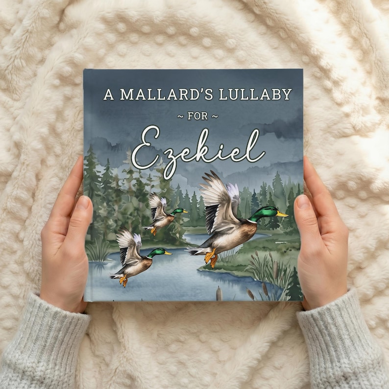 Może przedstawiać: Kwadratowa książka w twardej oprawie zatytułowana A Mallard's Lullaby For Ezekiel. Okładka zawiera akwarelową ilustrację trzech krzyż&oacute;wek lecących nad niebieską rzeką i zielonym lasem pod ciemnym, zachmurzonym niebem z białym napisem.