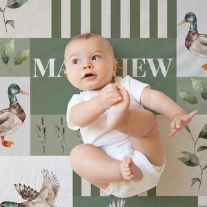 May include: A baby in a white onesie lies on a patterned blanket. The blanket features watercolor illustrations of ducks, leaves, and cattails, with the name "MATHEW" in white block letters against a green background.