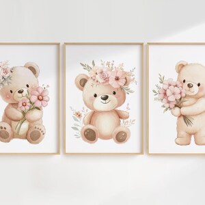May include: Set of three framed watercolor prints featuring adorable teddy bears. Each bear is depicted in soft, pastel colors, holding or adorned with pink flowers and floral accents. The prints are set against a white background.