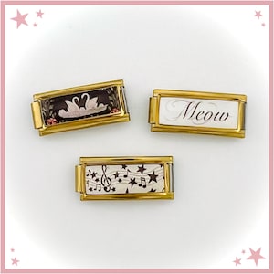 Puede incluir: Tres colgantes rectangulares dorados. Uno presenta dos cisnes blancos sobre un fondo negro con detalles florales. Otro colgante tiene la palabra "Meow" en escritura cursiva sobre un fondo blanco. El tercero muestra notas musicales y estrellas.