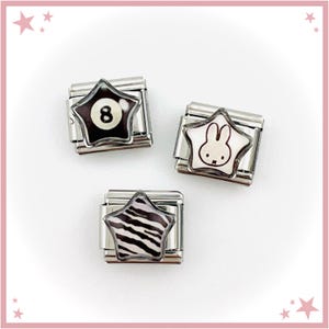May include: Three silver charm links with star-shaped designs. One has a black background with a white eight ball, another a white bunny outline, and the last a black and white zebra pattern. These charms are likely for a bracelet or necklace.