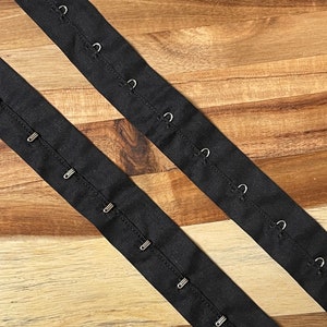 May include: Two black fabric strips with metal hooks and eyes for sewing onto garments. The strips are laid out on a wooden surface.