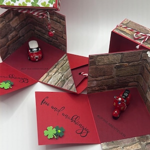 Könnte beinhalten: Eine rot-weiße Geschenkbox mit Kleeblatt- und Marienkäfermuster. Die Box öffnet sich und zeigt rote Tafeln mit Miniaturfahrzeugen und Ziegelstein-Akzenten. Text enthält "Zum grossen Tag" und "Auf ins Abenteuer".