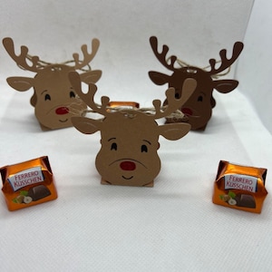 Könnte beinhalten: Drei braune Papier-Rentier-Geschenkboxen mit Bindfadenbögen. Zwei Boxen sind leer und eine Box enthält eine Schokoladen Süßigkeit, die in Goldfolie verpackt ist mit dem Text "Ferrero Küsschen".