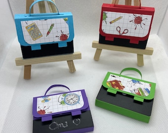 May include: Four miniature paper bag-shaped boxes with different colored handles and designs. The boxes are on small wooden easels. The boxes have a black chalkboard surface on the front. One box has a blue handle and a design of a calculator, pencil, and paint splatters. Another box has a red handle and a design of a pencil, scissors, and paint splatters. A third box has a green handle and a design of a book, pencil, and paint splatters. The fourth box has a purple handle and a design of a clock, butterfly, and paint splatters. The box with the purple handle has the name "Emma" written on the chalkboard.