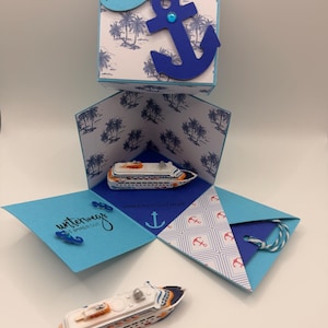 Könnte beinhalten: Eine nautische Pop-up-Box mit blauem Anker und Palmenmuster. Die Box öffnet sich und zeigt ein Miniatur-Kreuzfahrtschiff. Text enthält "Alles an Bord" und "Unterwegs ist immer gut."