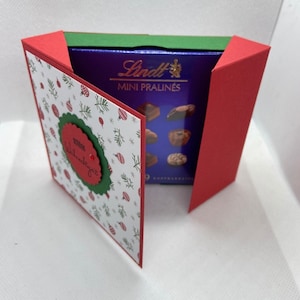 Peut inclure: Une carte de Noël rouge et verte avec un fond blanc et une petite boîte de mini pralines Lindt à l'intérieur. La carte a un design festif avec des ornements, des flocons de neige et une couronne avec le mot "Frohe Weihnachten" au centre.