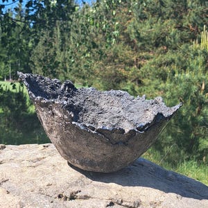 Op de afbeelding: Een handgemaakte, donkergrijze, getextureerde kom met een unieke, ongelijke rand. De kom staat op een grote, lichtgekleurde rots, met een achtergrond van groene bomen en een blauwe lucht. Deze decoratieve kom is perfect voor het bewaren van kleine items.
