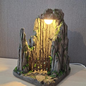 Könnte beinhalten: Eine dekorative Lampe in Form einer felsigen Berghöhle mit strukturierter grauer Oberfläche, grünen Moosflecken und einem kleinen braunen Holztor am Eingang. Eine warm leuchtende Glühbirne ist oben in der Felsformation angebracht.
