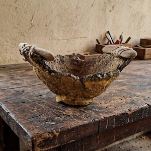 Könnte beinhalten: Eine texturierte, goldbraune Schale mit einer rauen, organischen Außenfläche. Die Schale verfügt über zwei glatte, helle Holzgriffe, die mit dünner, heller Schnur, die um das Holz und den Schalenrand gewickelt ist, an den Seiten befestigt sind.