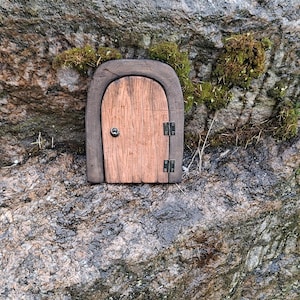 Puede incluir: Una pequeña puerta de madera arqueada con un panel de grano vertical marrón claro, un marco marrón oscuro, dos bisagras de metal negro en el lado derecho y un pequeño pomo redondo negro en el lado izquierdo, sobre una superficie de roca gris.