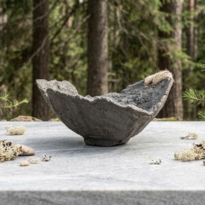May include: A dark gray, textured bowl with an irregular, jagged rim and a small base. The bowl features a rough, porous surface finish and a small piece of light-colored, fibrous twine attached to one side of the upper edge of the vessel.