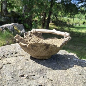 May include: A small, hand-formed bowl with a rough, textured, tan-coloured surface. The bowl features an irregular, jagged rim and a single, light-coloured wooden twig handle attached to the sides with thin, natural-coloured twine.