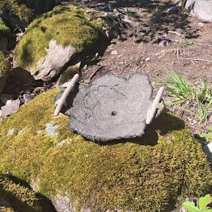 Puede incluir: Un marcador de piedra gris y desgastada con dos asas de madera, apoyado sobre una roca cubierta de musgo. El área circundante presenta más rocas musgosas y parches de tierra, creando una escena natural al aire libre.