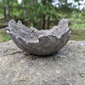 Puede incluir: Un pequeño objeto en forma de cuenco con una textura gris oscuro, rugosa e irregular. La superficie parece porosa y escarpada, similar a la piedra natural o al hormigón fundido. El borde es dentado e irregular, creando una cuenca profunda.