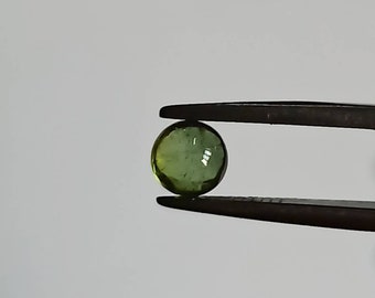 Puede incluir: Una piedra preciosa redonda y verde sostenida por unas pinzas. La piedra preciosa tiene una forma ligeramente irregular y una apariencia translúcida.