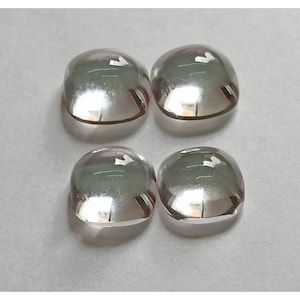 May include: Four clear, faceted, cushion-shaped gemstones with a slight pink hue. The gemstones are arranged in a square pattern on a white surface.