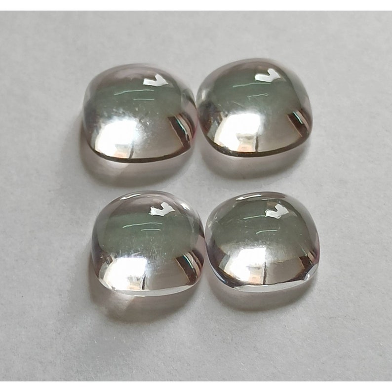 May include: Four clear, faceted, cushion-shaped gemstones with a slight pink hue. The gemstones are arranged in a square pattern on a white surface.