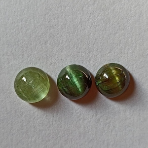 May include: Three green gemstone cabochons with a chatoyant effect. The cabochons are round and have a smooth, polished surface.
