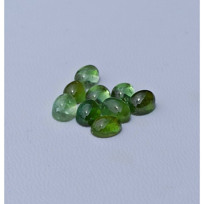 May include: A group of ten oval-shaped green gemstones, some with a slight yellow hue, are scattered on a white surface.