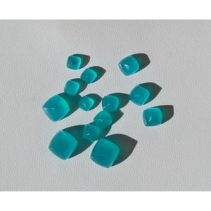 May include: A collection of teal blue gemstone cabochons, cut into a square shape with rounded edges. The gemstones are scattered on a white surface.