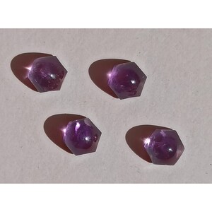 May include: Four hexagonal amethyst gemstones are arranged on a light background. Each gem is a deep purple colour and casts a shadow. The facets of the stones catch the light, creating a sparkling effect. These gemstones are ideal for jewellery making.