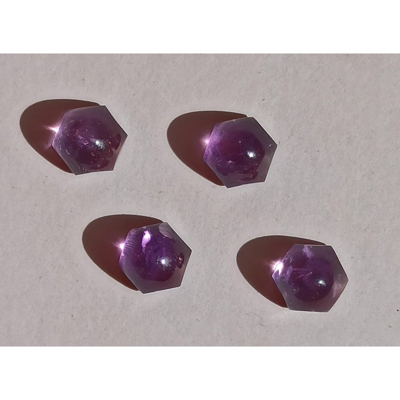 May include: Four hexagonal amethyst gemstones are arranged on a light background. Each gem is a deep purple colour and casts a shadow. The facets of the stones catch the light, creating a sparkling effect. These gemstones are ideal for jewellery making.