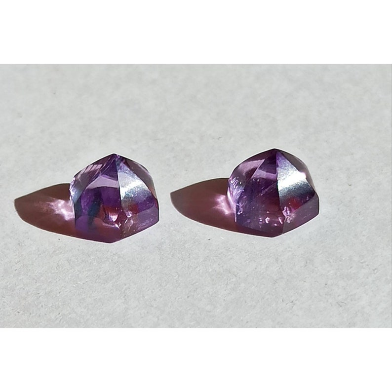 May include: Two faceted amethyst gemstones with a triangular shape. The gems are a deep purple colour and cast shadows on a white surface. The facets catch the light, creating a sparkling effect.