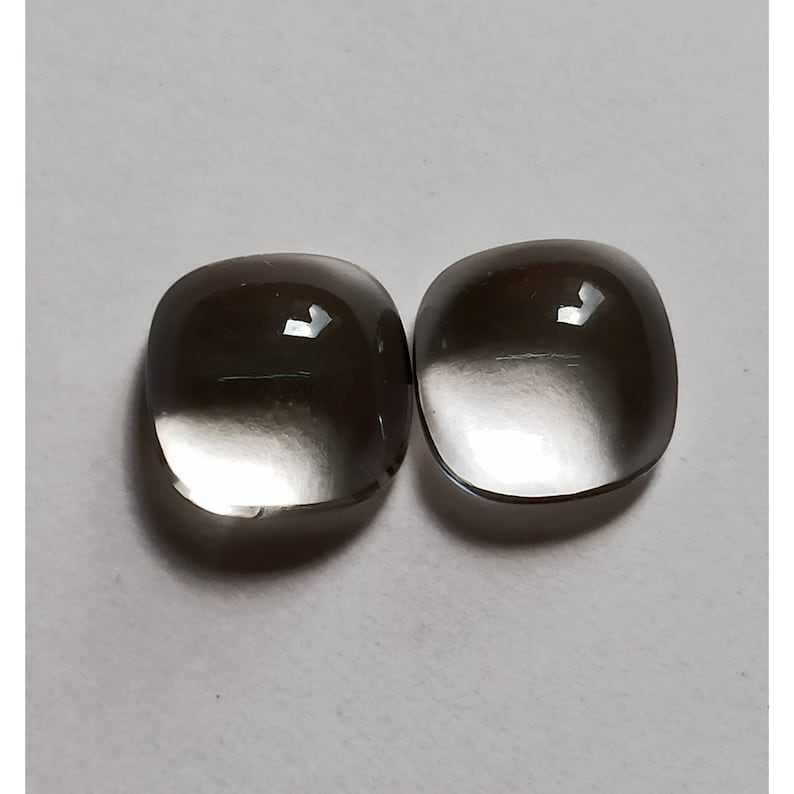 May include: Two clear, faceted, square-shaped gemstones with a smoky gray color gradient. The stones are smooth and polished.