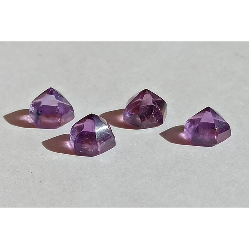 May include: Four faceted amethyst gemstones in a deep purple hue. Each gem is cut into a geometric shape, reflecting light and casting shadows. The gems are arranged on a light-coloured surface.