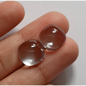 May include: Two clear, round, cabochon gemstones with a slight iridescent sheen. The stones are a light gray color with a hint of pink.