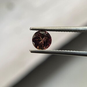 May include: A round, faceted gemstone with a reddish-brown color. The gemstone is held by tweezers.