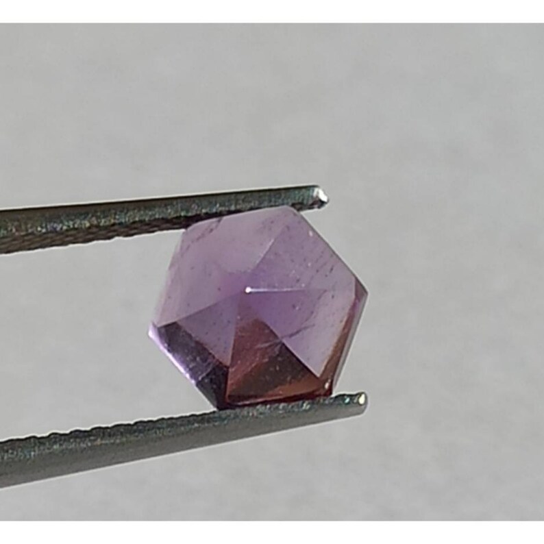 May include: A faceted, hexagonal gemstone held by tweezers. The gem has a deep purple hue with lighter purple facets. The tweezers are silver-coloured, and the background is a neutral grey.