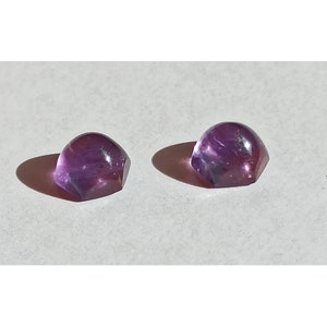 May include: Two faceted, dome-shaped amethyst gemstones. The gems are a deep purple colour with lighter purple and white inclusions. The stones are on a white surface, casting shadows.